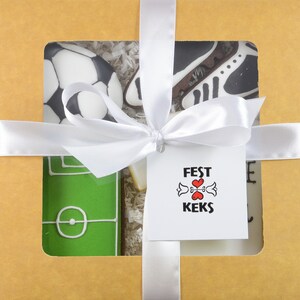 May include: A gift box with a white ribbon and tag. The box contains decorated cookies shaped like a soccer ball, soccer cleats, and a soccer field. The tag reads "FEST KEKS" with a heart design.