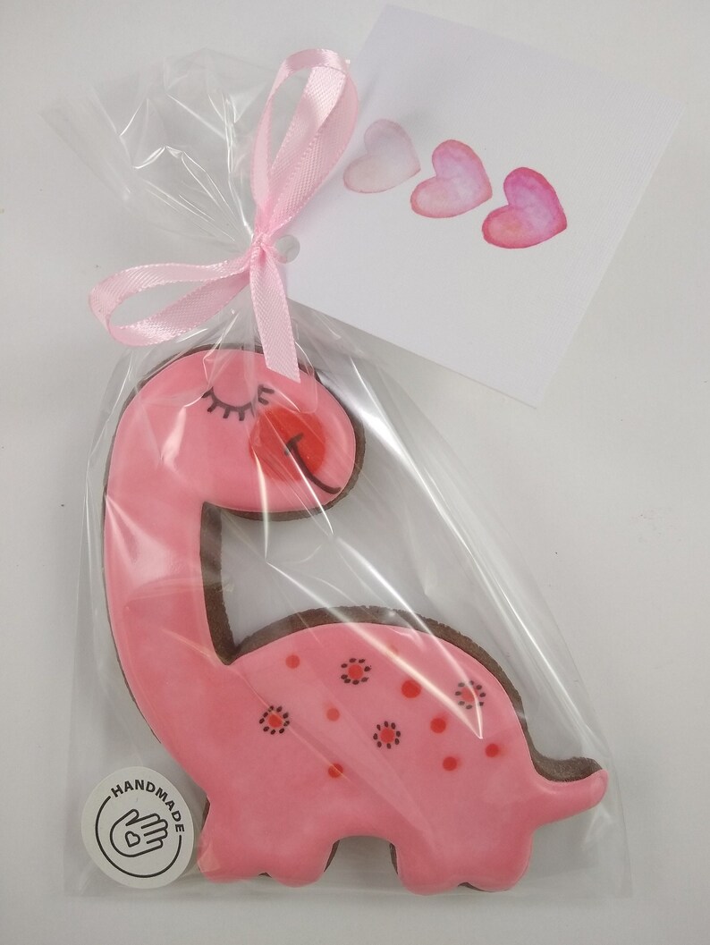 Op de afbeelding: Roze dinosauruskoekje met rode stippen en een handgetekend hartje aan de zijkant. Het koekje is verpakt in een doorzichtige plastic zak met een roze lint en een kaartje met de tekst "Handgemaakt" met een hartje.