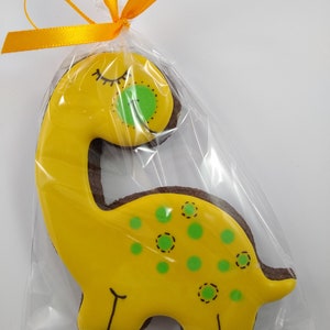 Op de afbeelding: Een gele dinosauruskoekje met groene stippen en een gesloten oog, versierd met zwarte glazuur, verpakt in een doorzichtige plastic zak met een geel lint.