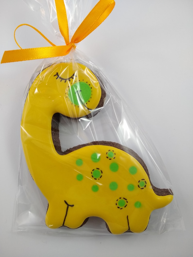 Op de afbeelding: Een gele dinosauruskoekje met groene stippen en een gesloten oog, versierd met zwarte glazuur, verpakt in een doorzichtige plastic zak met een geel lint.