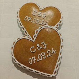 May include: Two heart-shaped gingerbread cookies decorated with white icing. The cookies are decorated with the initials "C & J" and the date "07.09.24".