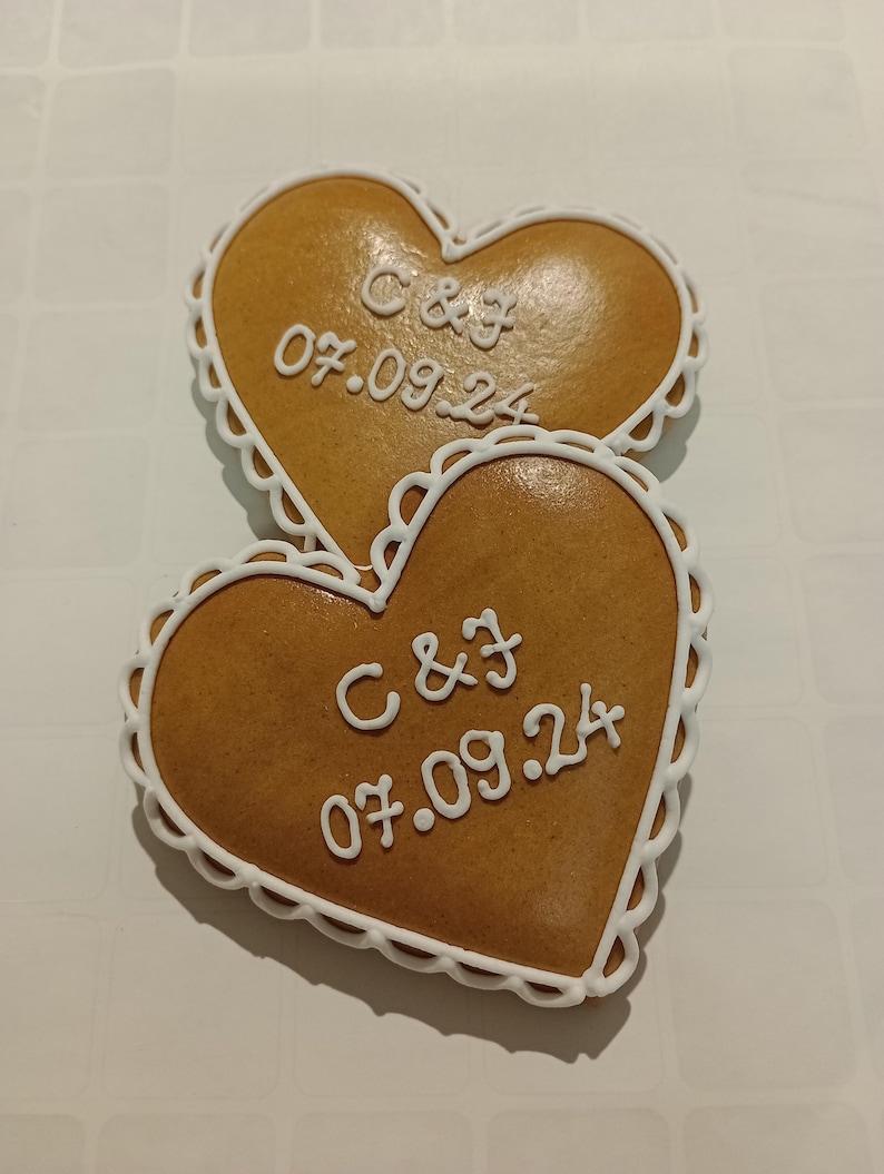 May include: Two heart-shaped gingerbread cookies decorated with white icing. The cookies are decorated with the initials "C & J" and the date "07.09.24".