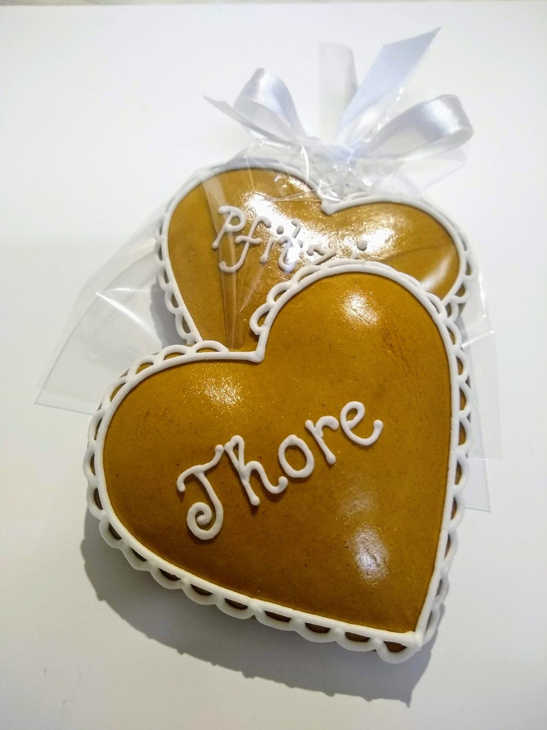 May include: Two heart-shaped gingerbread cookies decorated with white icing. The cookies are packaged in a clear plastic bag with a white ribbon. One cookie is decorated with the word "Fritz" and the other with the word "Thore".