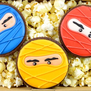 May include: Three decorated sugar cookies with ninja faces. The cookies are yellow, blue, and red. The cookies are surrounded by popcorn.