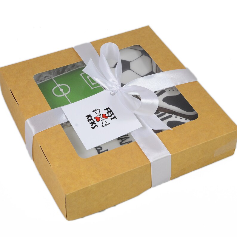 May include: A square, brown cardboard box with a clear window, tied with a white satin ribbon. Inside are soccer-themed treats, including a soccer ball and a soccer field design. A tag with the text "Fest Keks" is attached.