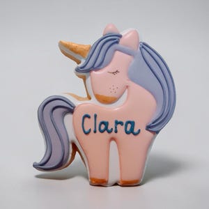 May include: A whimsical unicorn-shaped cookie with a pink body, lavender mane and tail, and a golden horn. The name "Clara" is written in blue script across the body. The unicorn has closed eyes and is set against a white background.