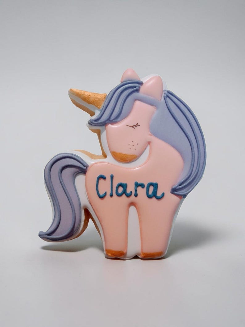 May include: A whimsical unicorn-shaped cookie with a pink body, lavender mane and tail, and a golden horn. The name "Clara" is written in blue script across the body. The unicorn has closed eyes and is set against a white background.