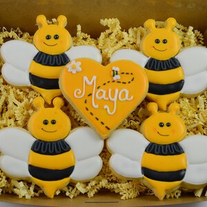 Könnte beinhalten: Eine Geschenkbox gefüllt mit gelben und schwarzen Bienen-Kekse. Die Kekse sind mit weißen Flügeln und schwarzen Streifen verziert. Ein herzförmiger Keks in der Mitte sagt "Maya".
