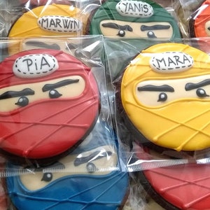 May include: Assortment of decorated sugar cookies with ninja faces. The cookies are red, yellow, green, and blue. Each cookie has a name written on it: Pia, Mara, Yanis, and Marvin.