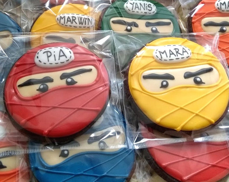 May include: Assortment of decorated sugar cookies with ninja faces. The cookies are red, yellow, green, and blue. Each cookie has a name written on it: Pia, Mara, Yanis, and Marvin.