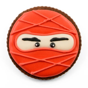 May include: A round chocolate cookie decorated with red icing and a white ninja mask with black eyes. The icing has a criss-cross pattern.