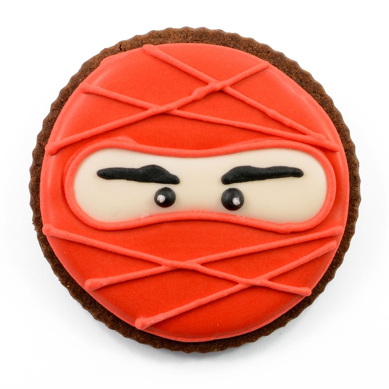 May include: A round chocolate cookie decorated with red icing and a white ninja mask with black eyes. The icing has a criss-cross pattern.
