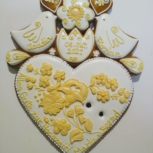May include: A white gingerbread heart with yellow floral designs and the text "Alles Gute zur Hochzeit" and "Marlis & Bernd". Two white doves with the names "Claudia" and "Florian" are on top of the heart, along with a yellow flower and a white egg with the date "08. Okt. 2027".
