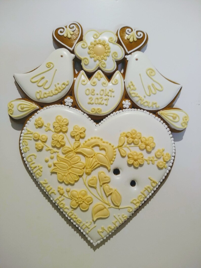May include: A white gingerbread heart with yellow floral designs and the text "Alles Gute zur Hochzeit" and "Marlis & Bernd". Two white doves with the names "Claudia" and "Florian" are on top of the heart, along with a yellow flower and a white egg with the date "08. Okt. 2027".