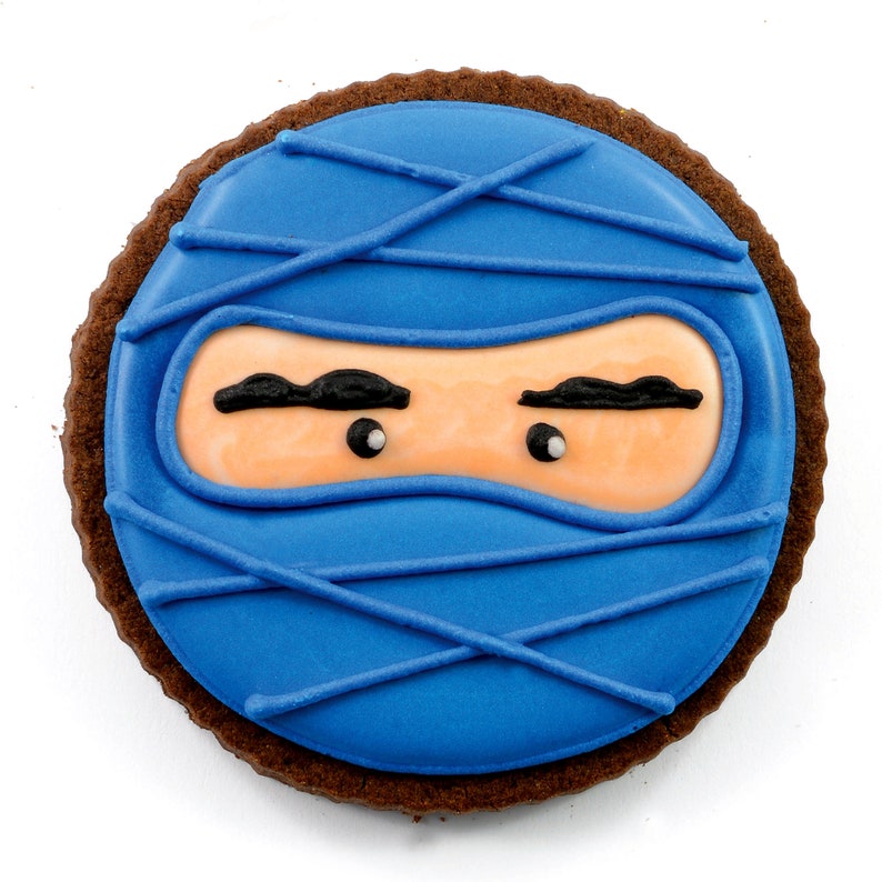 May include: A round chocolate cookie decorated with blue icing and a ninja face. The ninja has black eyebrows and white eyes.