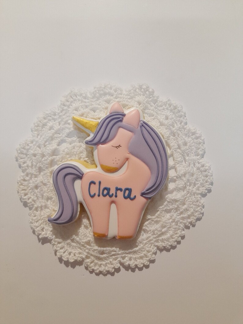 May include: A unicorn-shaped cookie with pink, purple, and gold details, resting on a white, lacy doily. The cookie has the name "Clara" written in blue script. The unicorn has closed eyes and a golden horn.