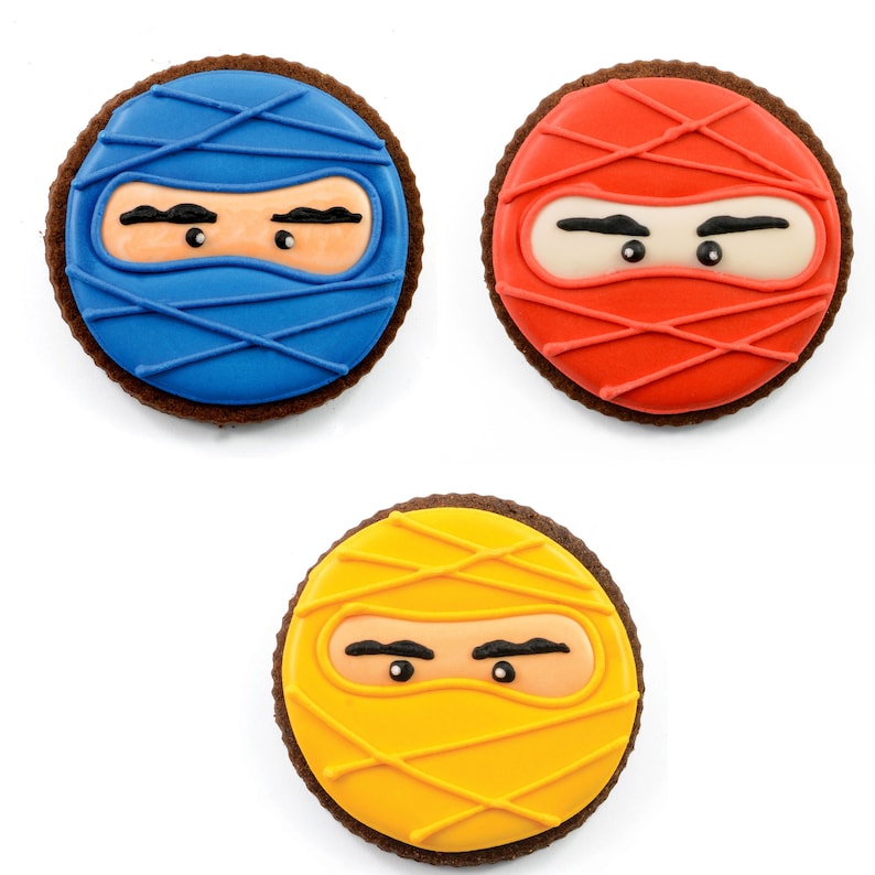 May include: Three decorated sugar cookies shaped like ninja faces. The cookies are brown with colorful icing. One cookie is blue, one is red, and one is yellow.