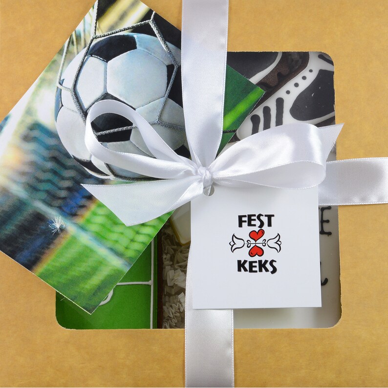 May include: A gift box tied with a white satin ribbon. Inside, a soccer-themed card and a tag that reads "FEST KEKS" with a heart and floral design. The box is a light brown color.