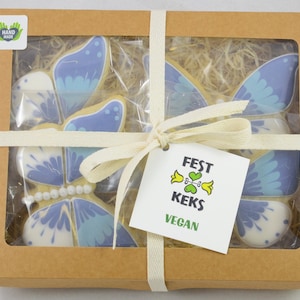 Op de afbeelding: Een bruine kartonnen doos met een transparant venster dat vier blauwe en witte vlinderkoekjes laat zien. De doos is vastgebonden met een wit lint en heeft een label met de tekst "FEST KEKS VEGAN".