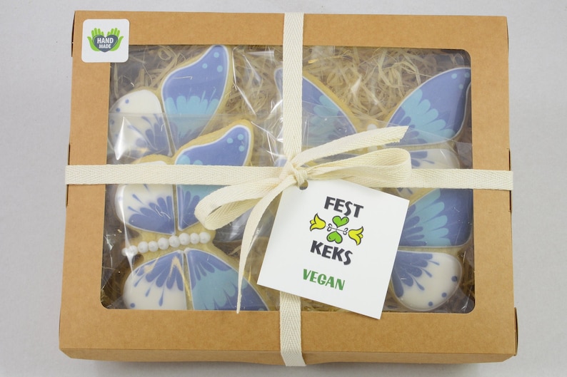 Op de afbeelding: Een bruine kartonnen doos met een transparant venster dat vier blauwe en witte vlinderkoekjes laat zien. De doos is vastgebonden met een wit lint en heeft een label met de tekst "FEST KEKS VEGAN".