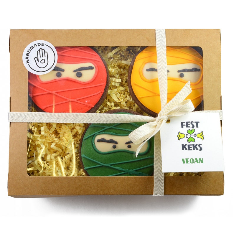 May include: A gift box with a clear window showing three round cookies decorated with ninja faces. The cookies are red, yellow, and green. The box is tied with a white ribbon and has a tag that says "FEST KEKS VEGAN".