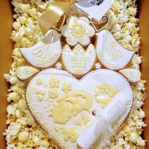 May include: A white heart-shaped gingerbread cookie with yellow floral designs and the text "Alles Gute zur Hochzeit" in a scroll. The cookie is surrounded by popcorn and a white ribbon. The date "08.04.2023" is written on a smaller cookie.