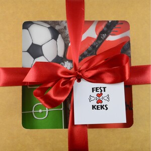 May include: A gift box wrapped in brown paper and tied with a red satin ribbon. The box features cutouts revealing soccer-themed cookies: a soccer ball, a soccer cleat, and a soccer field. A tag with the text "FEST KEKS" is attached.
