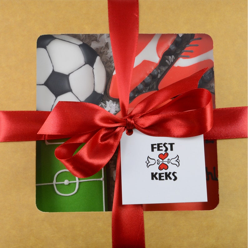 May include: A gift box wrapped in brown paper and tied with a red satin ribbon. The box features cutouts revealing soccer-themed cookies: a soccer ball, a soccer cleat, and a soccer field. A tag with the text "FEST KEKS" is attached.