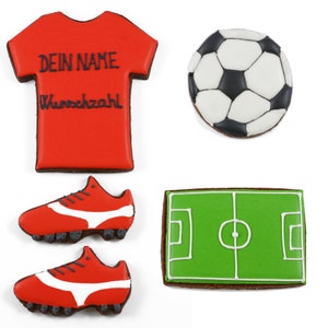 May include: A collection of decorated cookies in the shape of soccer-themed items. Includes a red jersey with the text "DEIN NAME Wunschzahl", a soccer ball, two red cleats with white accents, and a green field.