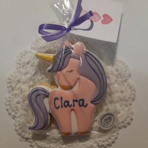 May include: A unicorn-shaped cookie with pink frosting, purple hair, and a golden horn, packaged in clear cellophane and tied with a purple ribbon. The cookie has the name "Clara" written on it. A small card with pink hearts is attached.