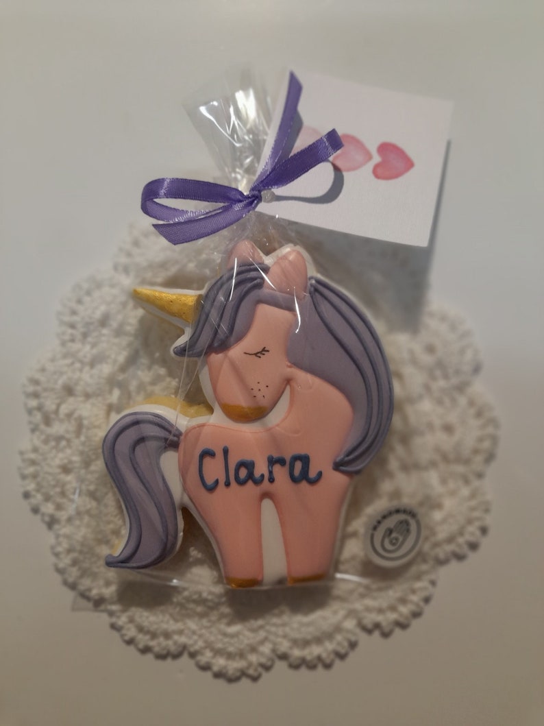 May include: A unicorn-shaped cookie with pink frosting, purple hair, and a golden horn, packaged in clear cellophane and tied with a purple ribbon. The cookie has the name "Clara" written on it. A small card with pink hearts is attached.