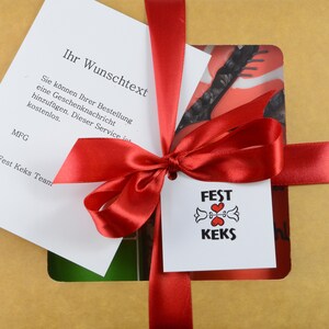 May include: A gift box tied with a red satin ribbon and bow. A white tag with the words "FEST KEKS" and a heart design is attached. A card with German text is visible, along with a decorative insert.