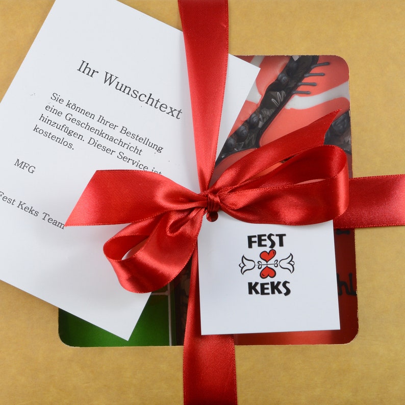 May include: A gift box tied with a red satin ribbon and bow. A white tag with the words "FEST KEKS" and a heart design is attached. A card with German text is visible, along with a decorative insert.