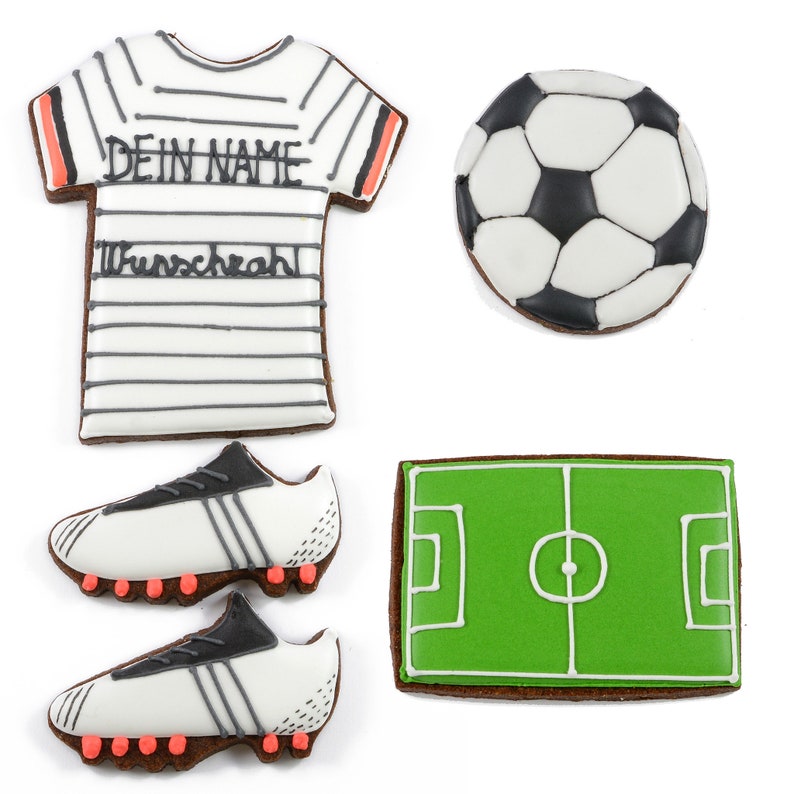 May include: A collection of soccer-themed cookies. Includes a white and black soccer ball, a green field, a white jersey with text, and two white soccer cleats with black and red accents. The jersey has the text "DEIN NAME" and "Wunschzahl".