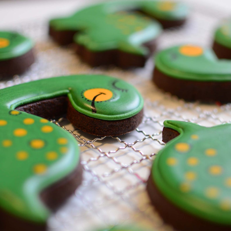 Op de afbeelding: Een close-up van verschillende groene en gele gedecoreerde koekjes op een draadrek. De koekjes zijn gevormd als dinosaurussen en hebben gele stippen en zwarte ogen.