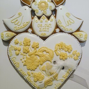 May include: A white gingerbread heart with yellow icing decorations and the text "Alles Gute zur Hochzeit" and "Marlis & Bert". Two white doves with yellow icing details and the names "Claudia" and "Florian" are above the heart. A yellow flower and two brown hearts are above the doves. The date "08. Okt. 2027" is written on a white egg-shaped gingerbread piece.