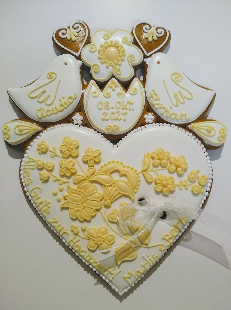 May include: A white gingerbread heart with yellow icing decorations and the text "Alles Gute zur Hochzeit" and "Marlis & Bert". Two white doves with yellow icing details and the names "Claudia" and "Florian" are above the heart. A yellow flower and two brown hearts are above the doves. The date "08. Okt. 2027" is written on a white egg-shaped gingerbread piece.