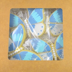 Op de afbeelding: Een doos gevuld met blauwe en witte vlinder-vormige koekjes. De koekjes zijn in een enkele laag gerangschikt en omgeven door wit tissuepapier.