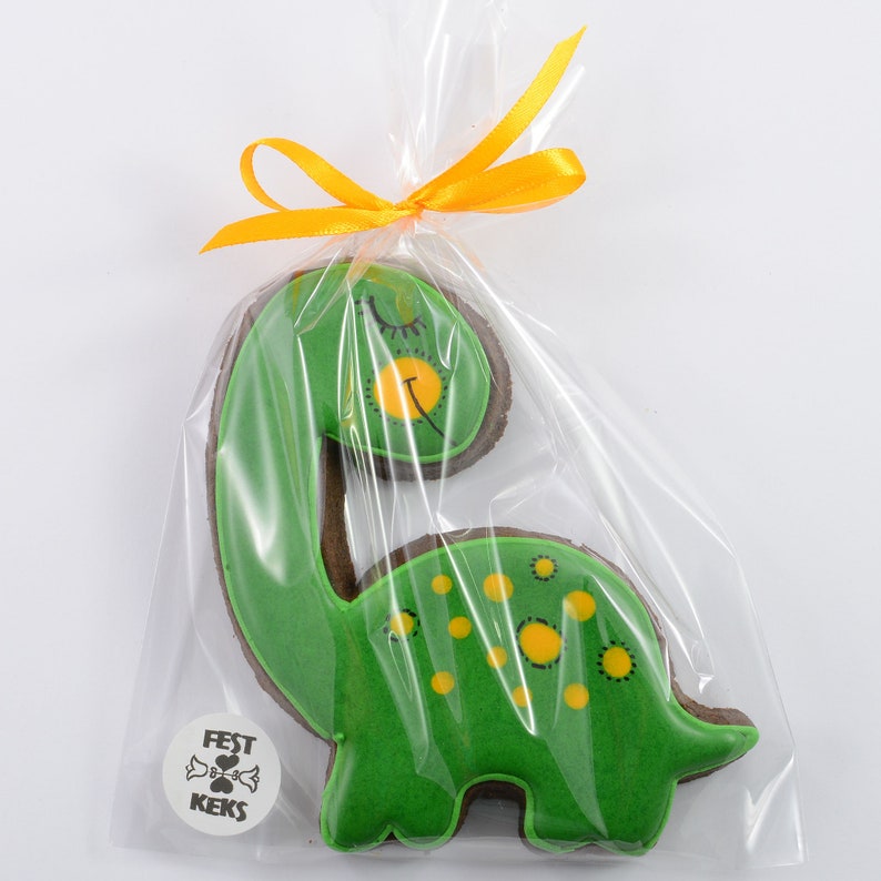 Op de afbeelding: Een groene en bruine dinosaurusvormige koek met gele stippen en een gesloten oog. De koek is verpakt in een doorzichtige plastic zak met een geel lint.