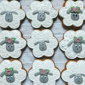 May include: Nine sheep-shaped cookies with white icing and gray sheep faces. Some cookies have pink rose and green leaf decorations. The cookies are arranged in a grid pattern, perfect for a spring or Easter treat.