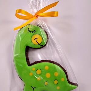 Op de afbeelding: Een groene dinosaurusvormige koek met gele stippen en een slapend gezicht, versierd met zwarte glazuur. De koek is verpakt in een doorzichtige plastic zak met een geel lint.