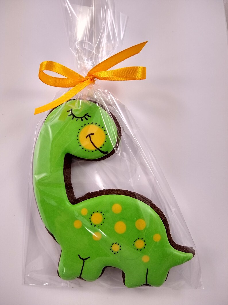 Op de afbeelding: Een groene dinosaurusvormige koek met gele stippen en een slapend gezicht, versierd met zwarte glazuur. De koek is verpakt in een doorzichtige plastic zak met een geel lint.