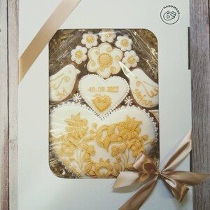 May include: A white gift box with a gold ribbon and a clear window showing a set of gingerbread cookies decorated with gold icing. The cookies are shaped like hearts, birds, and flowers. The date "4.09.2022" is written on one of the cookies. The box has a "HANDMADE" label with a hand icon.