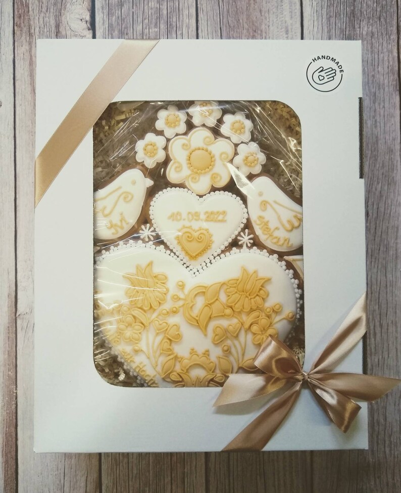 May include: A white gift box with a gold ribbon and a clear window showing a set of gingerbread cookies decorated with gold icing. The cookies are shaped like hearts, birds, and flowers. The date "4.09.2022" is written on one of the cookies. The box has a "HANDMADE" label with a hand icon.