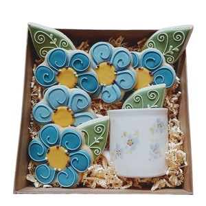 May include: A gift box containing flower-shaped cookies with blue icing and yellow centers, green leaf cookies, and a white mug with blue floral designs. The cookies are arranged in a brown box with shredded paper.
