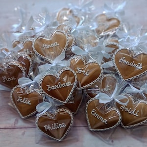 May include: A collection of heart-shaped gingerbread cookies, individually wrapped in clear cellophane bags with white ribbons. Each cookie is decorated with white icing and a name, including "Anna", "Max", "Felix", "Emma", "Hendrik", "Heidi", "Barbara", "Philine", "Toii", and "Robert".