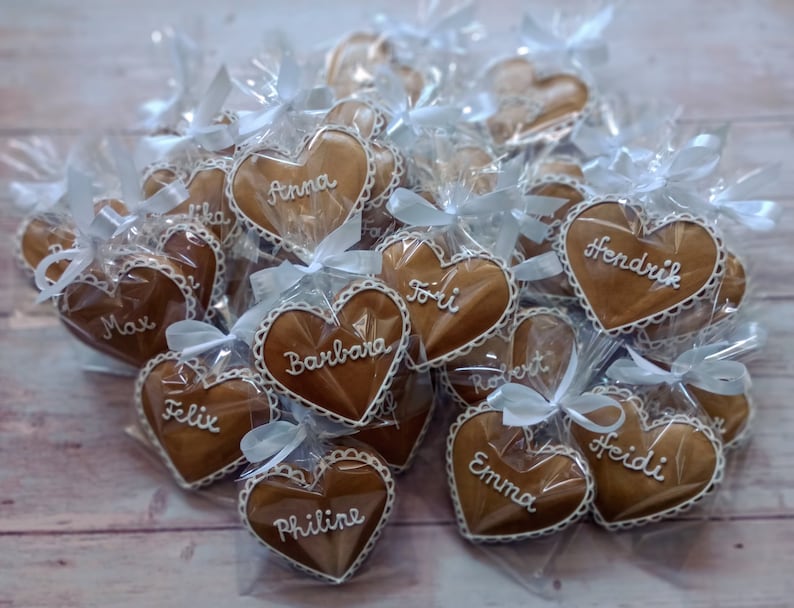 May include: A collection of heart-shaped gingerbread cookies, individually wrapped in clear cellophane bags with white ribbons. Each cookie is decorated with white icing and a name, including "Anna", "Max", "Felix", "Emma", "Hendrik", "Heidi", "Barbara", "Philine", "Toii", and "Robert".