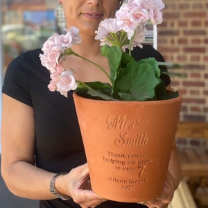 May include: A terracotta flower pot with a personalised message that reads "Mrs. Smith Thank you for helping me grow in year 7 Alison Moore 2022". The pot is filled with a pink geranium plant.