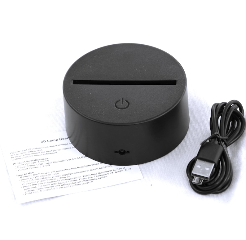 May include: A black 3D lamp base with a slot for an acrylic plate, a power button, and a USB cable. The base is round with a flat top and a slightly angled side. A user manual is included.