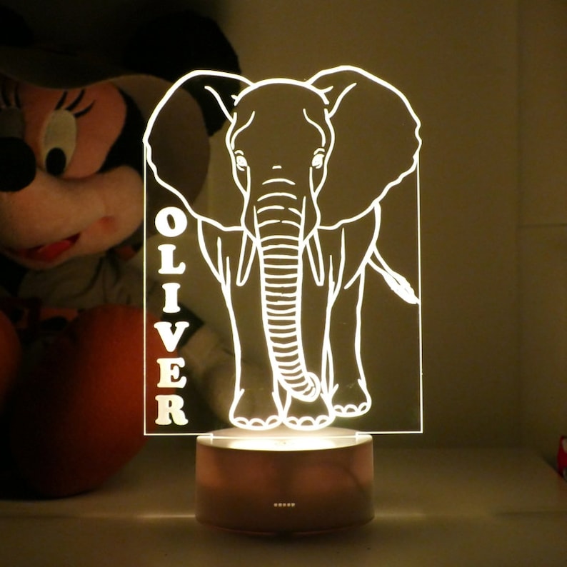 May include: A glowing LED night light featuring a white elephant design with the name "OLIVER" vertically displayed. The elephant is illuminated on a clear acrylic panel, set on a brown base. The light emits a warm glow.